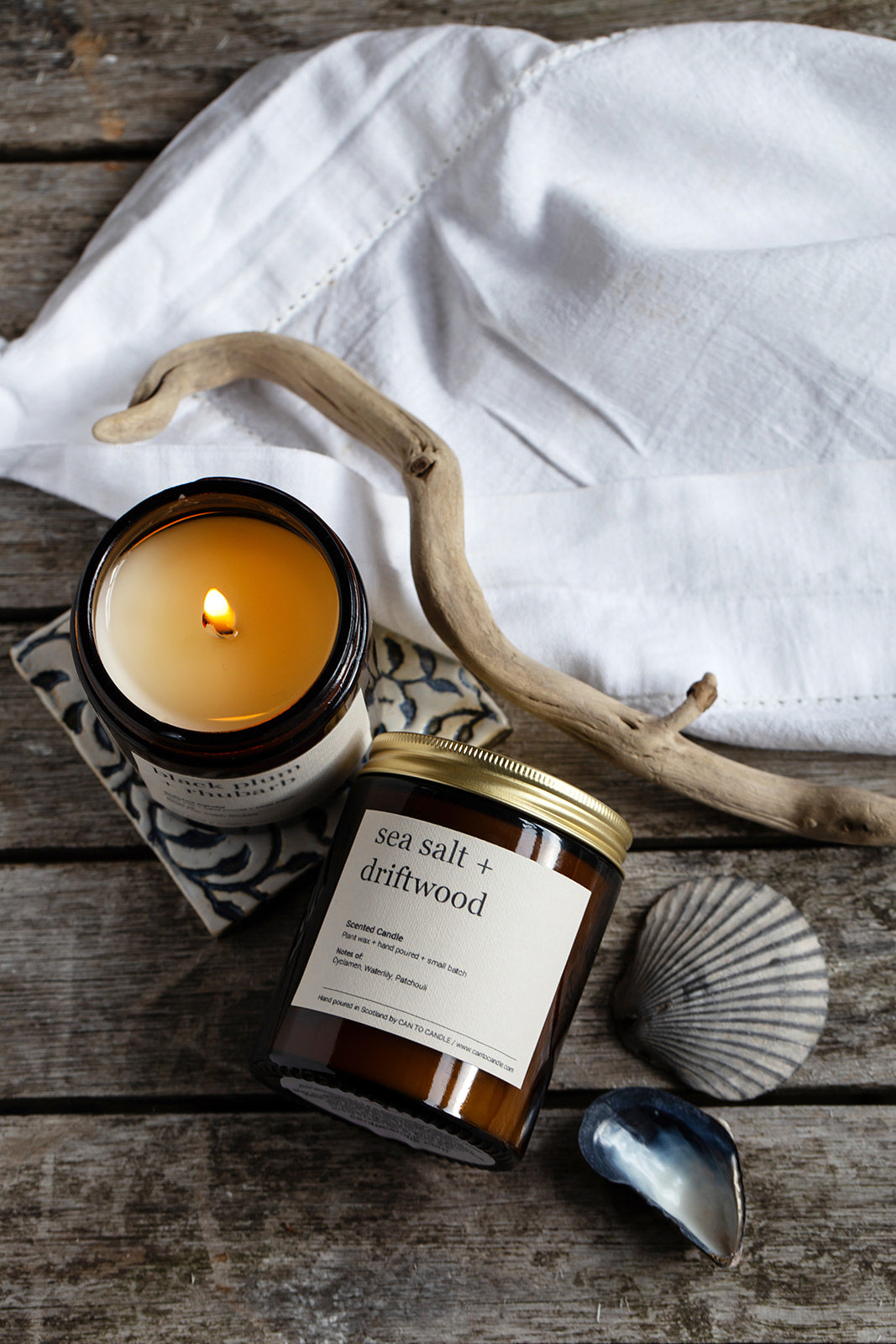 Candle by Kerry of Can to Candle and jar labeled 'sea salt & driftwood' on a wooden surface with a white cloth and shell in the background.