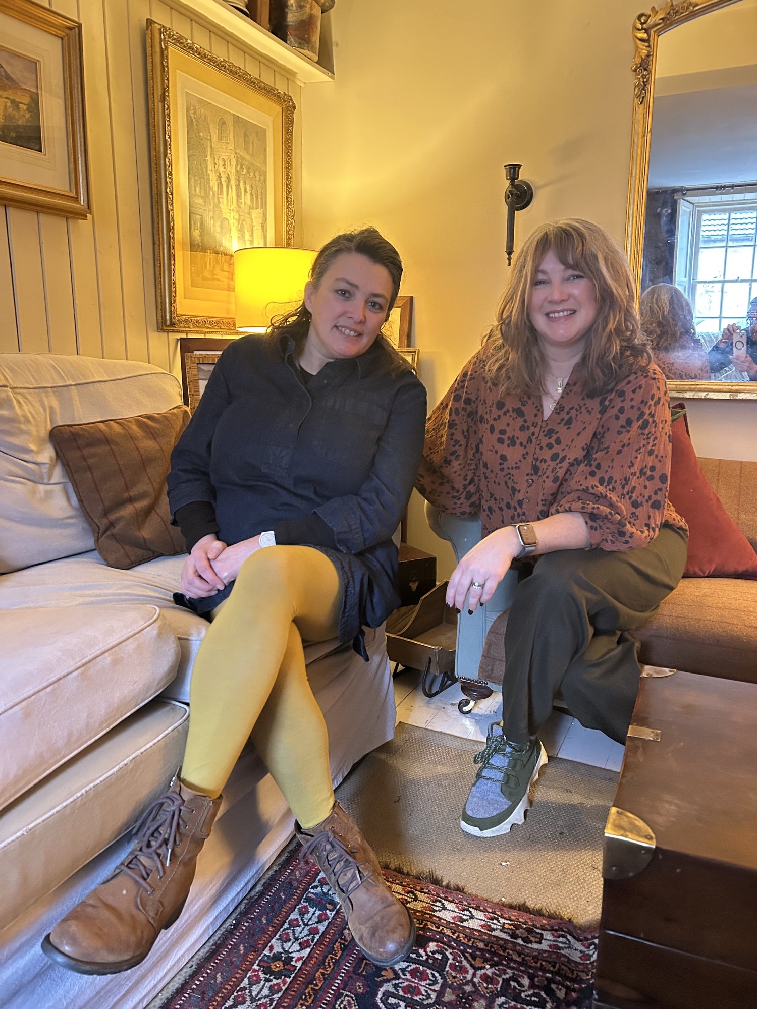 Two women. (Helen and Kerry) sitting on a couch in a cozy living room with framed pictures and a lamp.