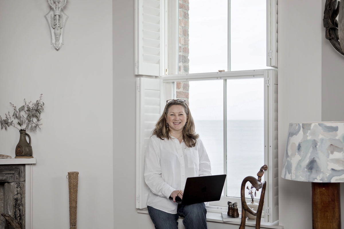 Woman (Helen of HR Digital Design) holding a laptop in a room with large windows and decorative elements.